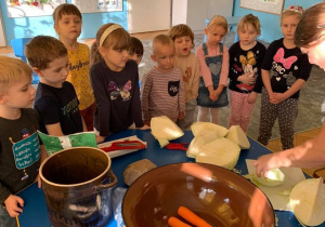 Na stole stoi garnek, miska z obraną marchewką, pałka, tarka oraz hebelki. Nauczycielka kroi na desce kapustę na małe kawałki. 10 dzieci obserwuje czynność.